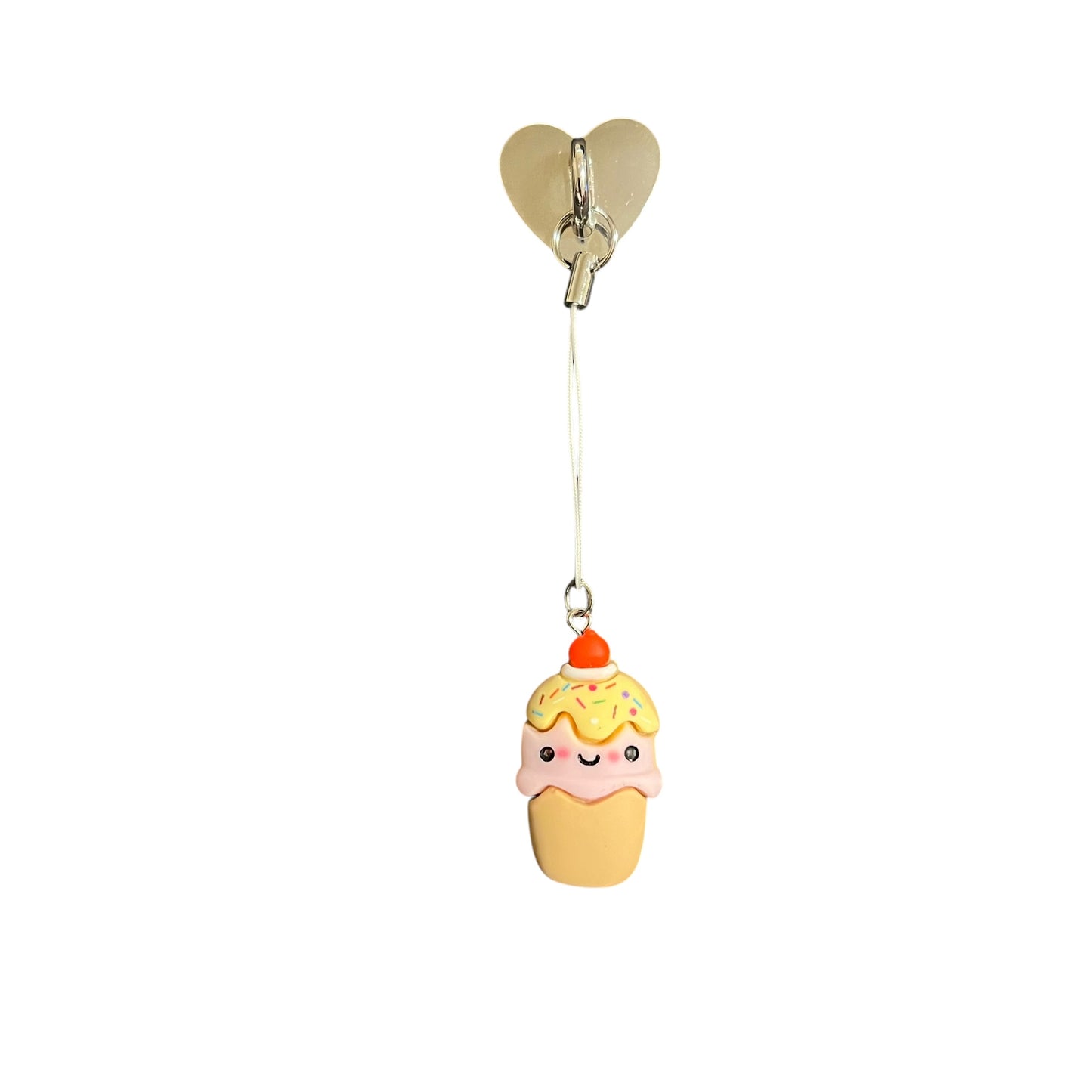 A handmade ice cream cone phone charm with a smiling face, rainbow sprinkles, and a cherry on top, attached to a gold-tone heart mount with a strap.