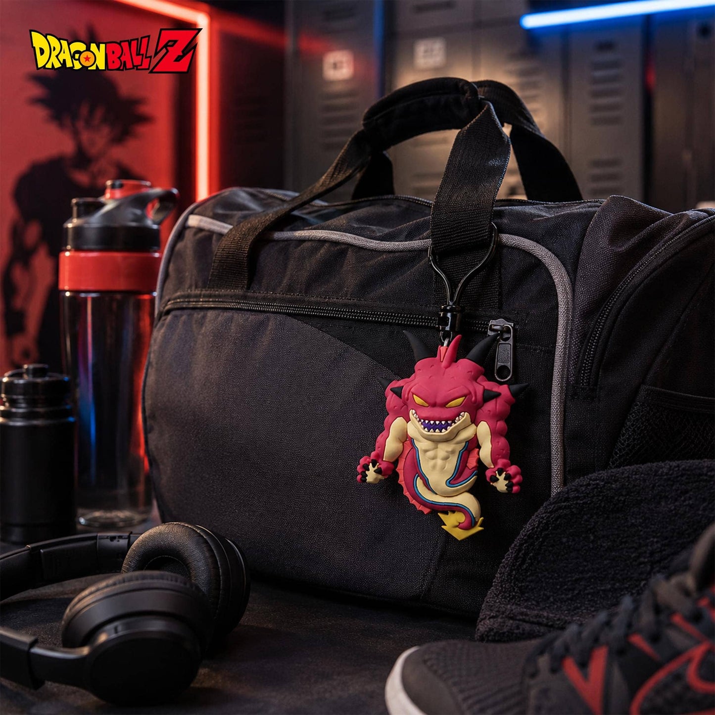 A black Dragon Ball Z themed gym duffel bag with a hanging pink Fused Ball character charm, styled on a dark table with a water bottle, headphones, and sneakers in a locker room setting.