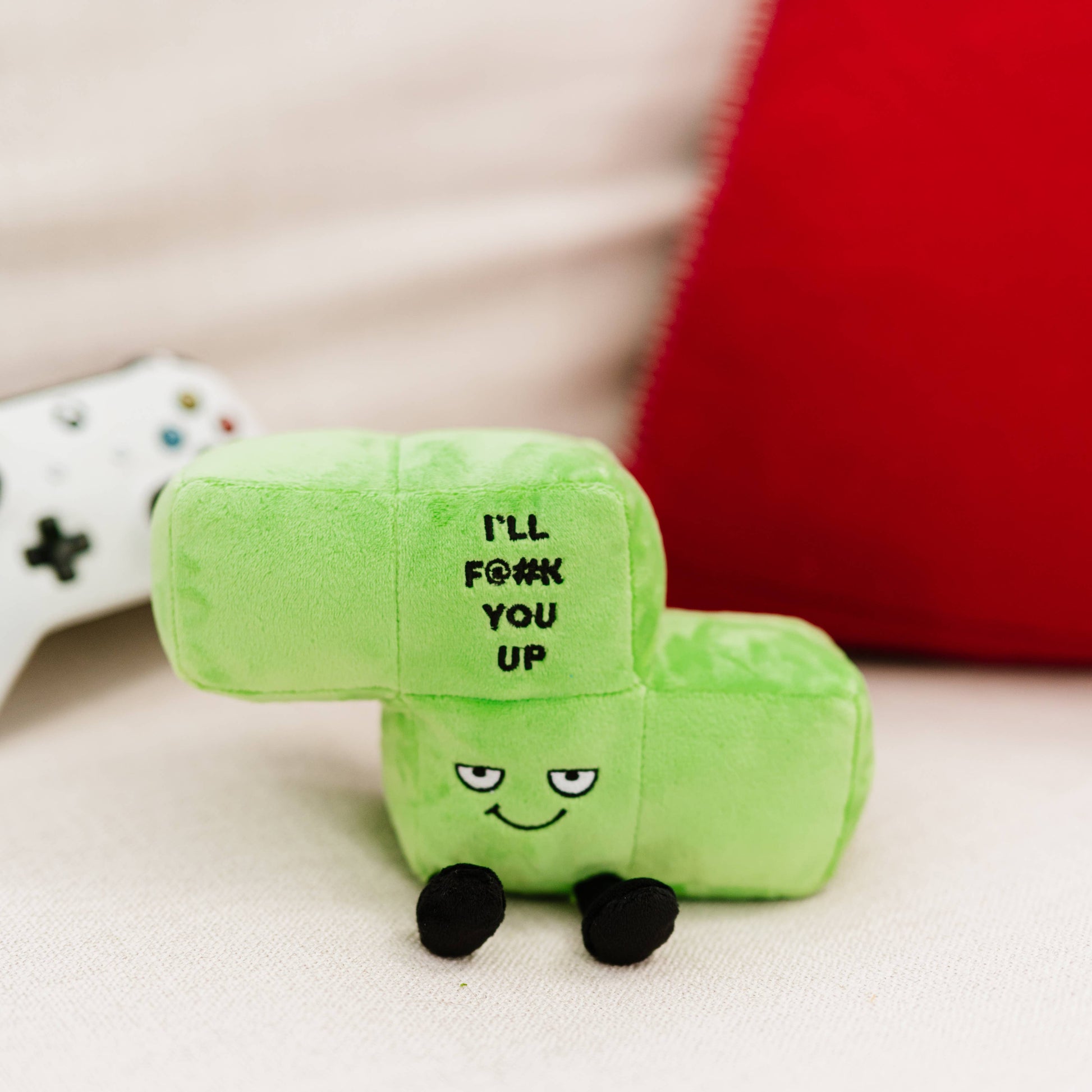 A green block-shaped plush toy with a cartoon face and the text 'I'LL F*#K YOU UP' on the front, photographed on a light fabric surface with a red cushion and part of a white game controller in the background.