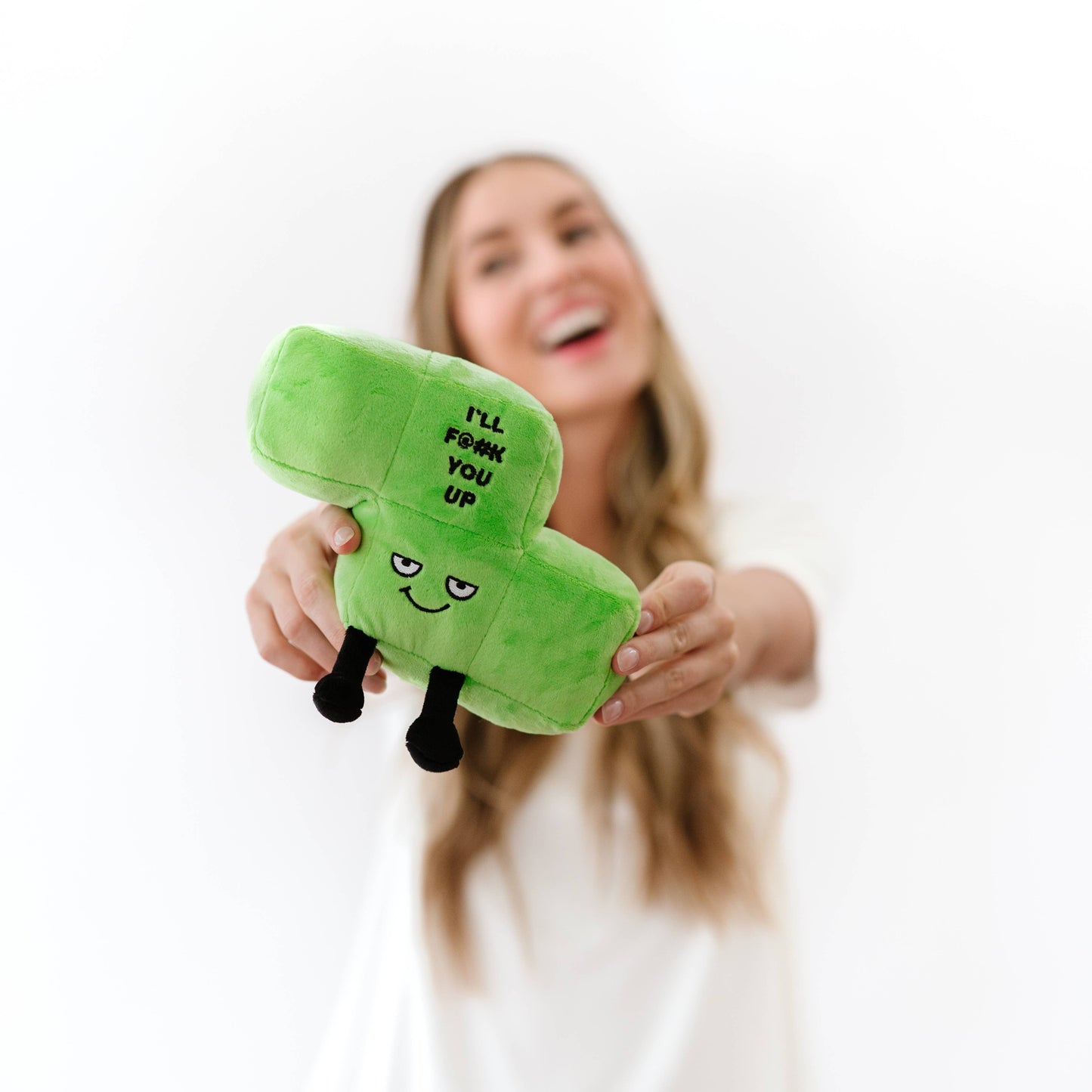 A woman holding a green plush toy shaped like a block with the text 'I'LL F*CK YOU UP' and a simple face, against a bright white background.