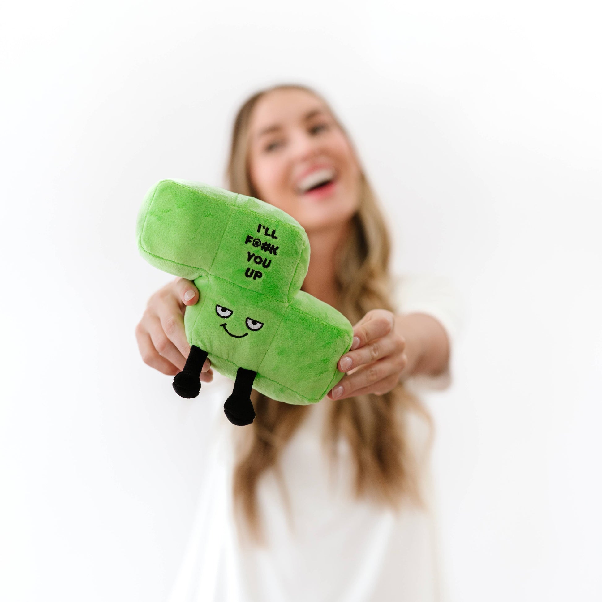 A woman holding a green plush toy shaped like a block with the text 'I'LL F*CK YOU UP' and a simple face, against a bright white background.