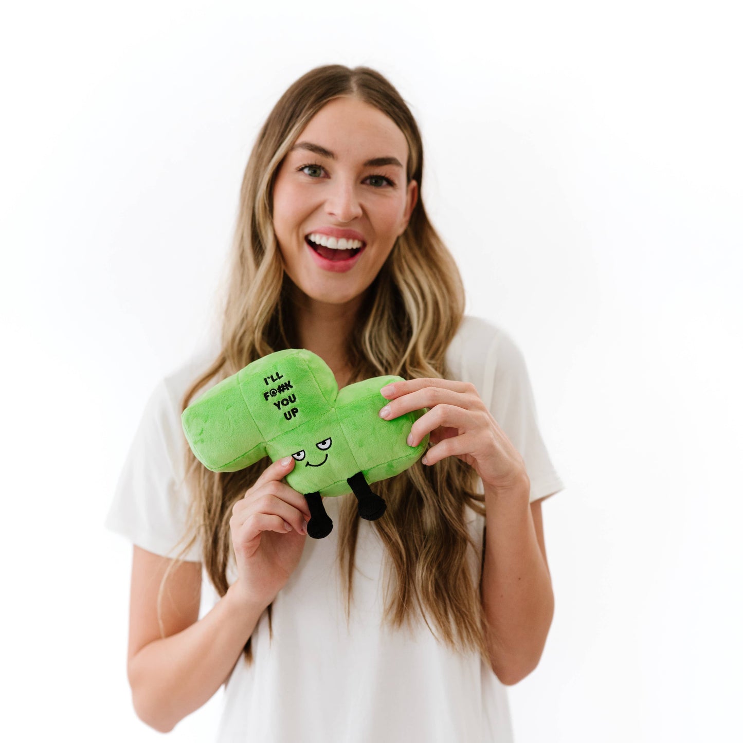 A smiling woman with long blonde hair holds a bright green plush pillow shaped like a slice of pizza. The pillow has a cartoon face and the text 'I'LL FROM YOU UP' printed on it.