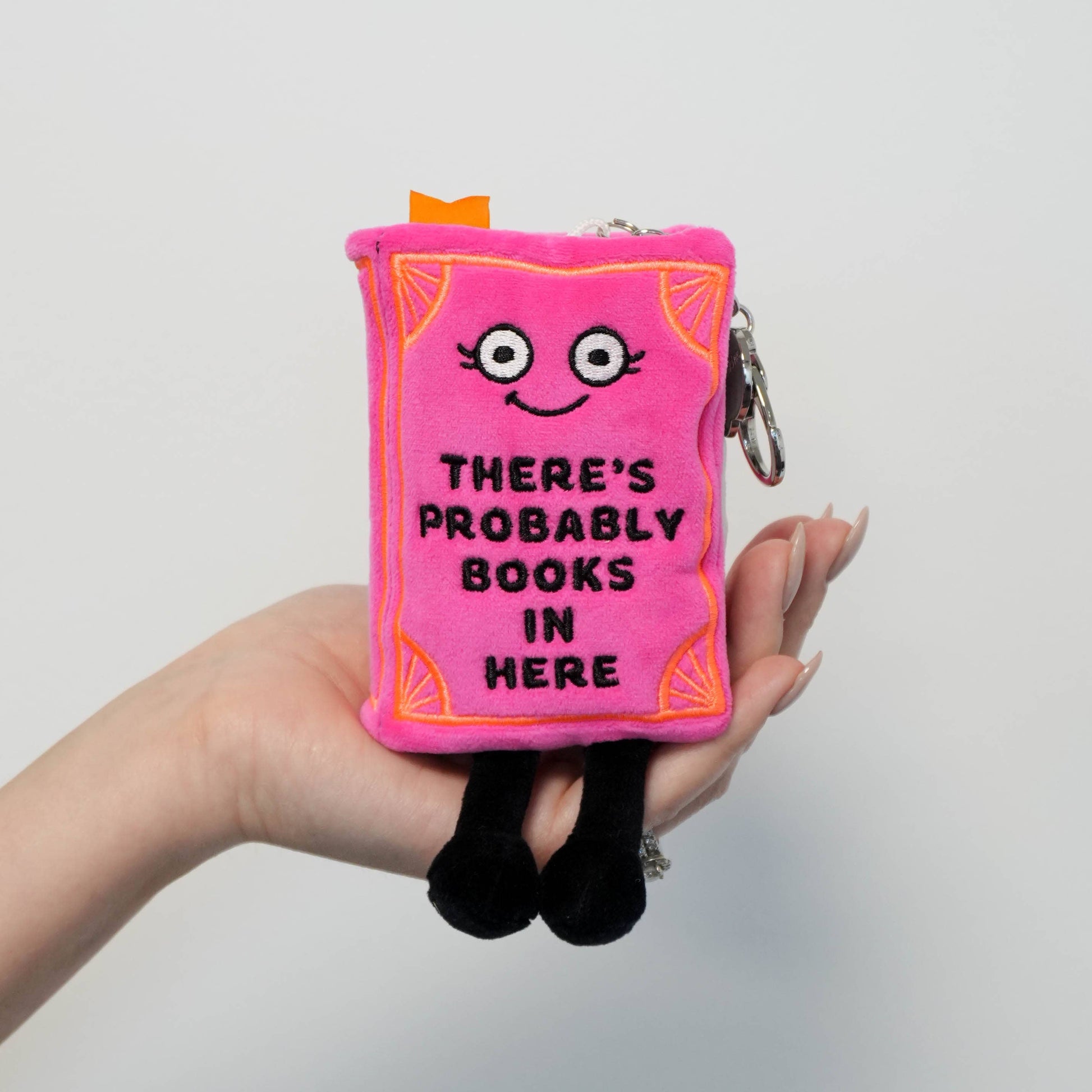 Pink plush book-shaped pouch with a face and text held by a hand against a light gray background